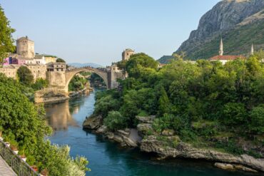 A picturesque view of the historic Stari Most bridge spanning the turquoise Neretva River, surrounded by lush greenery and historic architecture in Mostar, Bosnia and Herzegovina.